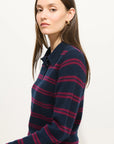 A woman with long brown hair wears the Alex Mill Alice Polo Cashmere, a luxury navy blue sweater with magenta stripes, and faces slightly to the side, looking at the camera against a plain white background.