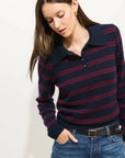 A woman with long brown hair is wearing the Alex Mill Alice Polo Cashmere sweater in navy with red stripes, blue jeans, and a black belt, standing against a light background and looking down with one hand in her pocket.