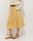 Model in a cream sweater tucked into the Alex Mill Maddie Skirt Linen—a loose, knee-length, garment-dyed skirt with pockets—styled with a brown belt and brown flat sandals, standing against a white background.