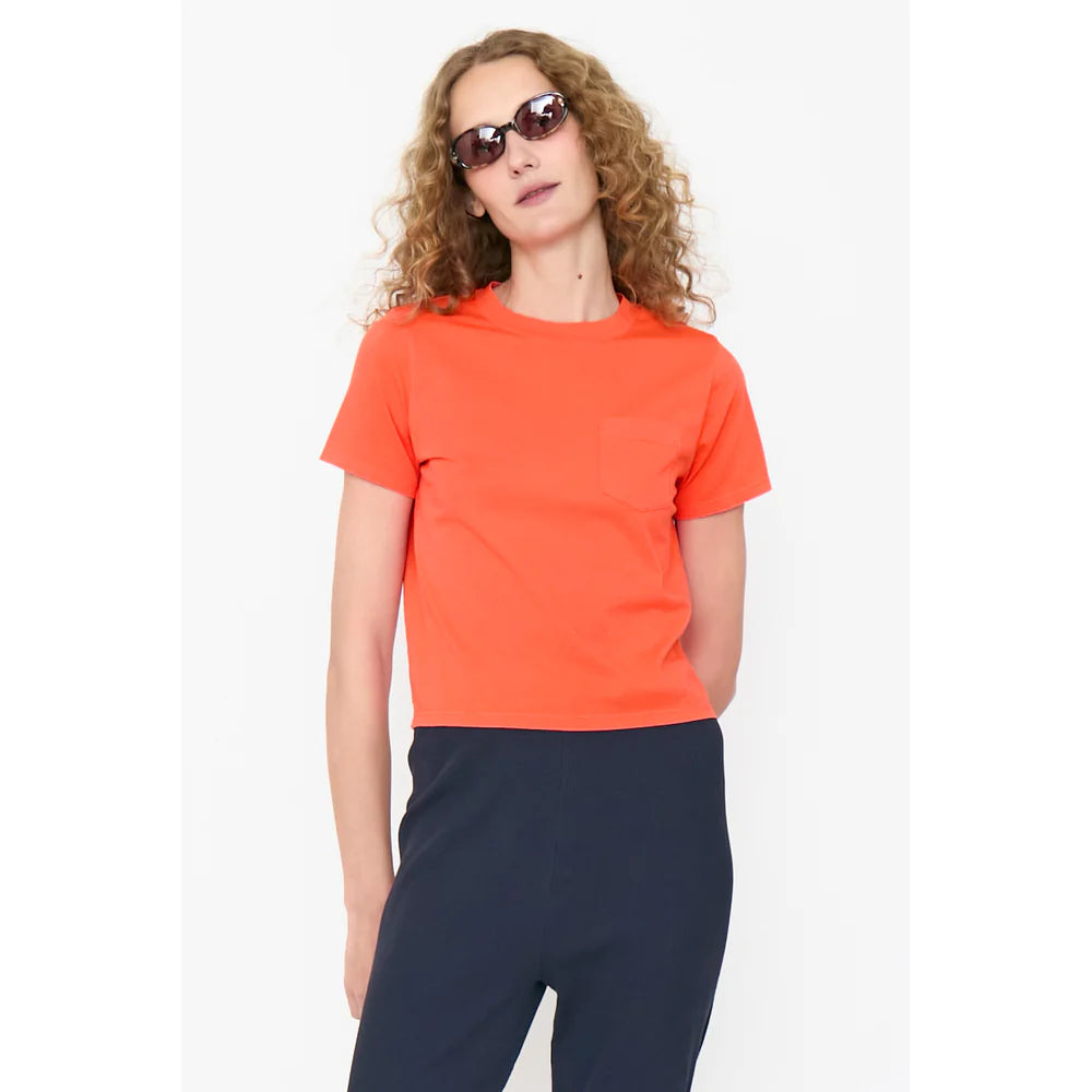 A person with curly hair in dark sunglasses, dark pants, and a short-sleeve orange cotton The Wynn Baby Tee by Kule stands against a plain white background.