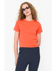 A person with curly hair in dark sunglasses, dark pants, and a short-sleeve orange cotton The Wynn Baby Tee by Kule stands against a plain white background.