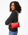 A woman with curly hair, wearing a black sweater and blue jeans, smiles while holding the Clare Vivier Wallet Clutch w/Tabs, crafted from Italian nappa leather with a gold chain strap. She stands against a white background, exuding elegance with her made-in-India style accessory.