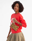 A woman with curly hair wears a red sweatshirt and beige pleated skirt, holding the Clare Vivier Wallet Clutch w/Tabs in Italian suede with a gold chain, standing against a plain white background.