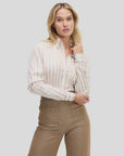 A woman with wavy blonde hair models the Weekend Shirt by Sarah Alexandra, LLC, featuring neutral stripes, tucked into high-waisted light brown pants. She poses against a plain background with one hand on her collar and the other on her hip.
