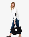 A woman wears the Kerri Rosenthal Mia Shirt Classic White with black elbow patches, blue jeans, and black flats, holding a large black tote bag with a white heart against a light background for a timeless wardrobe staple look.