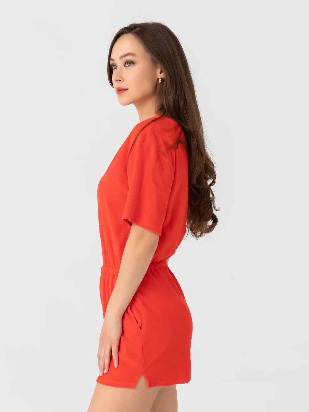 A woman with long brown hair, wearing the Xirena Evert Romper in bright red towel terry, stands sideways against a plain light gray background.