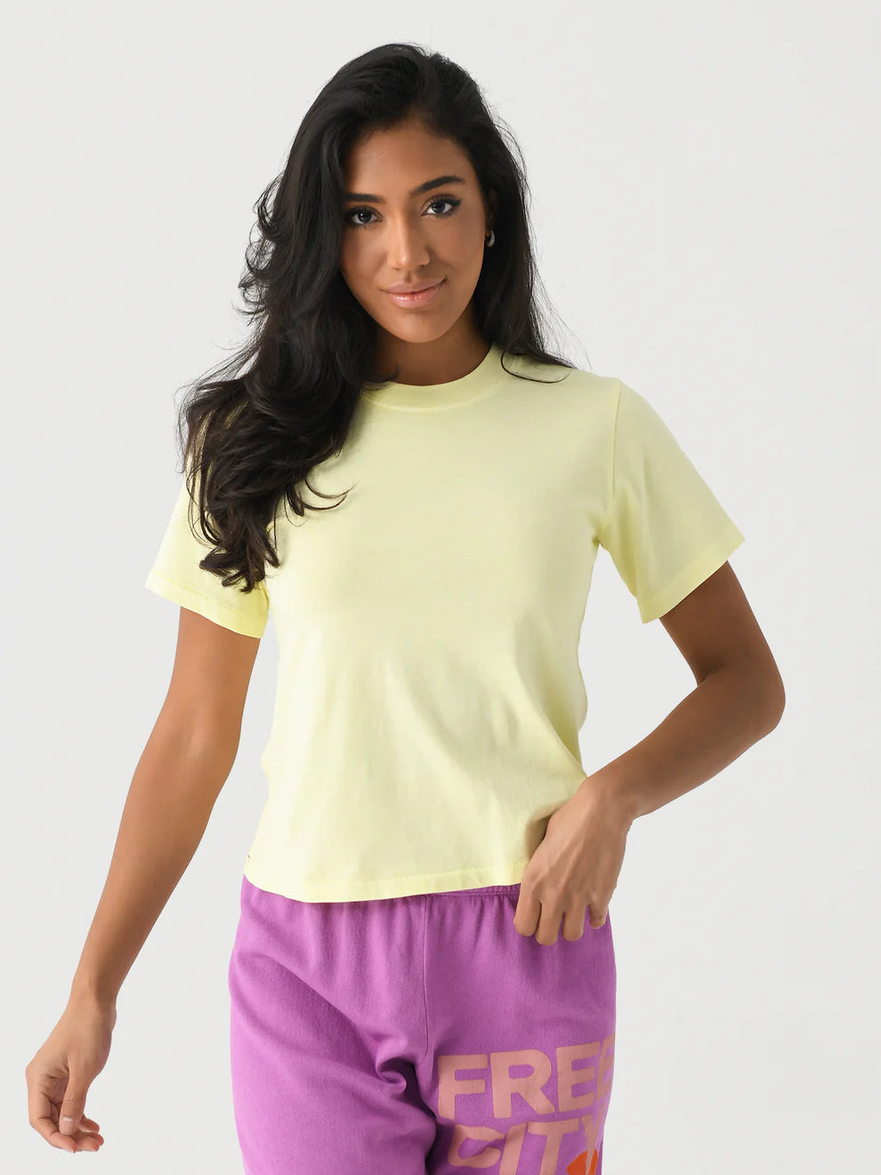 A woman with long dark hair wears the Xirena Georgy Tee in pale yellow, styled with purple “FREE CITY” shorts in orange and pink, standing against a white background.