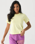 A woman with long dark hair wears the Xirena Georgy Tee in pale yellow, styled with purple “FREE CITY” shorts in orange and pink, standing against a white background.