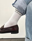 A person wearing light blue jeans and brown loafers has their leg crossed, revealing the Perfectwhitetee "You Are Perfect Sock" in white ribbed cotton with black text reading "you are perfect.