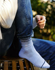 A person in jeans and a light sweater sits on a wooden stool, wearing Perfectwhitetee’s You Are Perfect Sock—vintage-inspired ribbed cotton socks with “you are perfect” near the top. Blurred greenery forms the backdrop.