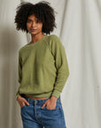 A person with short curly hair is wearing a green Ziggy Reverse Fleece LS Crew Sweatshirt from Perfectwhitetee, featuring exposed seam detailing, and blue jeans. They are standing in front of a plain light-colored backdrop with one hand in their pocket and the other relaxed by their side. The expression on their face appears calm.