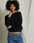 A person with curly hair poses against a plain backdrop, wearing a cozy essential: the Ziggy Reverse Fleece LS Crew Sweatshirt by Perfectwhitetee in black, paired with blue jeans. One hand rests on their chin while the other is crossed over their waist.