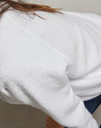 A close-up of a person wearing the Perfectwhitetee Ziggy Reverse LS Sweatshirt in textured white cotton fleece and blue jeans, leaning forward slightly. Only their upper body, arm, and hair are visible.