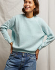 Wearing a Ziggy Reverse Fleece LS Crew Sweatshirt by Perfectwhitetee with exposed seam detailing and blue jeans, the person smiles with hands on hips against a light, textured curtain.