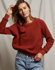 A person with long brown hair is wearing a Ziggy Reverse Fleece LS Crew Sweatshirt by Perfectwhitetee and blue jeans. They pose against a light fabric background, one hand touching their hair and the other in their pocket.