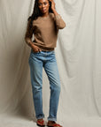 A woman poses before a beige fabric backdrop wearing the Perfectwhitetee Ziggy Reverse LS Sweatshirt in brown, paired with light blue jeans and red sandals. She has long curly hair, looking at the camera with one hand in her pocket and the other touching her hair.