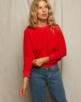 A woman with wavy blonde hair wears the Perfectwhitetee Ziggy Reverse Fleece LS Crew Sweatshirt in bright red and blue jeans, standing before a beige fabric backdrop, looking slightly to the side with one hand touching her sleeve.