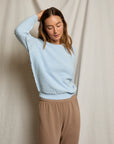 A woman stands before a light beige fabric backdrop, wearing the Perfectwhitetee Ziggy Reverse LS Sweatshirt in light blue and tan pants. She raises one hand to her head and looks down with a relaxed expression.