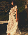 A woman stands on a sunlit beach, wearing the DOEN Zinnia Skirt—a sheer white lace two-piece with an asymmetrical hem and short sleeves—surrounded by rocks, greenery, and a bare tree in the background.
