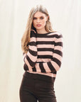 A person with long, straight blonde hair wears the Jumper 1 2 3 4/CR2 Cashmere Stripe Crew in pink and brown with black pants, standing against a plain, light background and touching their hair.