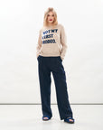 A woman poses confidently in the Rodeo Crew sweater by Jumper 1 2 3 4/CR2, featuring blue "NOT MY FIRST RODEO" text, paired with navy wide-leg trousers and blue sandals against a plain white background. Her hands are in her pockets.