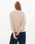 A person with long, straight hair stands facing away against a plain background, wearing the beige Rodeo Crew organic jumper by Jumper 1 2 3 4/CR2 and dark pants, hands behind their back, fingers loosely intertwined.