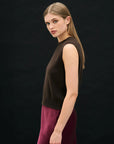 A woman with straight, light brown hair stands sideways against a plain black background, wearing the Jumper 1 2 3 4/CR2 Distressed Tank and maroon pants, calmly looking towards the camera.