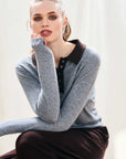 A woman with light hair pulled back wears a gray Jumper 1 2 3 4/CR2 Contrast Polo with a dark collar and brown pants, sitting with one arm bent and looking directly at the camera against a light background.