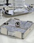 The Zodax Cocktail Napkin Holder Nickel, featuring a spherical weight, keeps blue and white napkins secure on your table. Perfect as a bar accessory, it sits elegantly beside a glass of water, silver tray, and decanter.