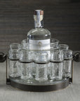 The Zodax Ten Shot Tequila Set w/Ice Bucket features a tall bottle at the center, surrounded by eight textured shot glasses, all arranged on a gray surface with a striped backdrop.