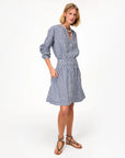 A woman stands against a white background, wearing the Clare Vivier Zoe Skirt—a blue and white linen gingham skirt with a smocked waist and pockets—paired with black studded sandals.