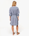 A woman with blonde hair faces away, wearing the Clare Vivier Zoe Skirt—a blue and white linen gingham piece with a smocked waist—paired with tan studded sandals, set against a plain white background.