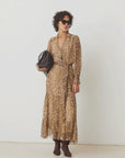 A woman with short curly hair, sunglasses, and brown boots models the SUNCOO Cosmos Dress—a loose-fit leopard print viscose dress—while holding a black handbag against a plain white background.