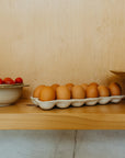 The Indaba Homestead Egg Holder displays brown eggs on a wooden shelf, serving as stylish storage and decor, placed beside a bowl of red cherry tomatoes against a light wood background.