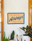 The Oxford Pennant "True Love Will Always Have It's Way" felt wall hanging features red and blue text, inspired by John Prine’s lyrics. Displayed above green plants and near wood-framed glass doors, it brightens any white wall.
