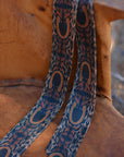 The Faire Western Harvest Twilly scarf features hand-drawn horseshoe and leafy vine patterns in brown and gold tones, displayed over a rustic brown leather chair.