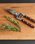 The Faire Thai Kitchen Garden Shears, featuring stainless steel blades and wooden handles, rest on a cutting board alongside fresh rosemary sprigs and sliced cherry tomatoes.