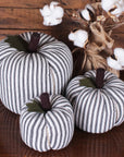 The Faire Pumpkin set features three black-and-white striped fabric pumpkins on a wooden surface beside burlap-wrapped cotton stems, creating charming farmhouse decor—perfect autumn ornaments.