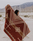 A person stands in a sandy landscape wrapped in the Achuar Blanket by Faire, with wind turbines and mountains in the background, creating a serene scene with boho decor vibes.
