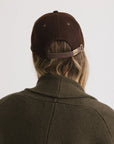 Someone with long hair is seen from behind wearing the Faire Aven Leather Ball Cap in dark brown, paired with an olive green sweater, set against a plain light background—a chic winter accessory.