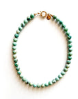 The Faire Galleria Necklace is a 17-inch gold-plated piece with round, light turquoise-green beaded stones and a gold clasp, displayed in a circle on a white background.