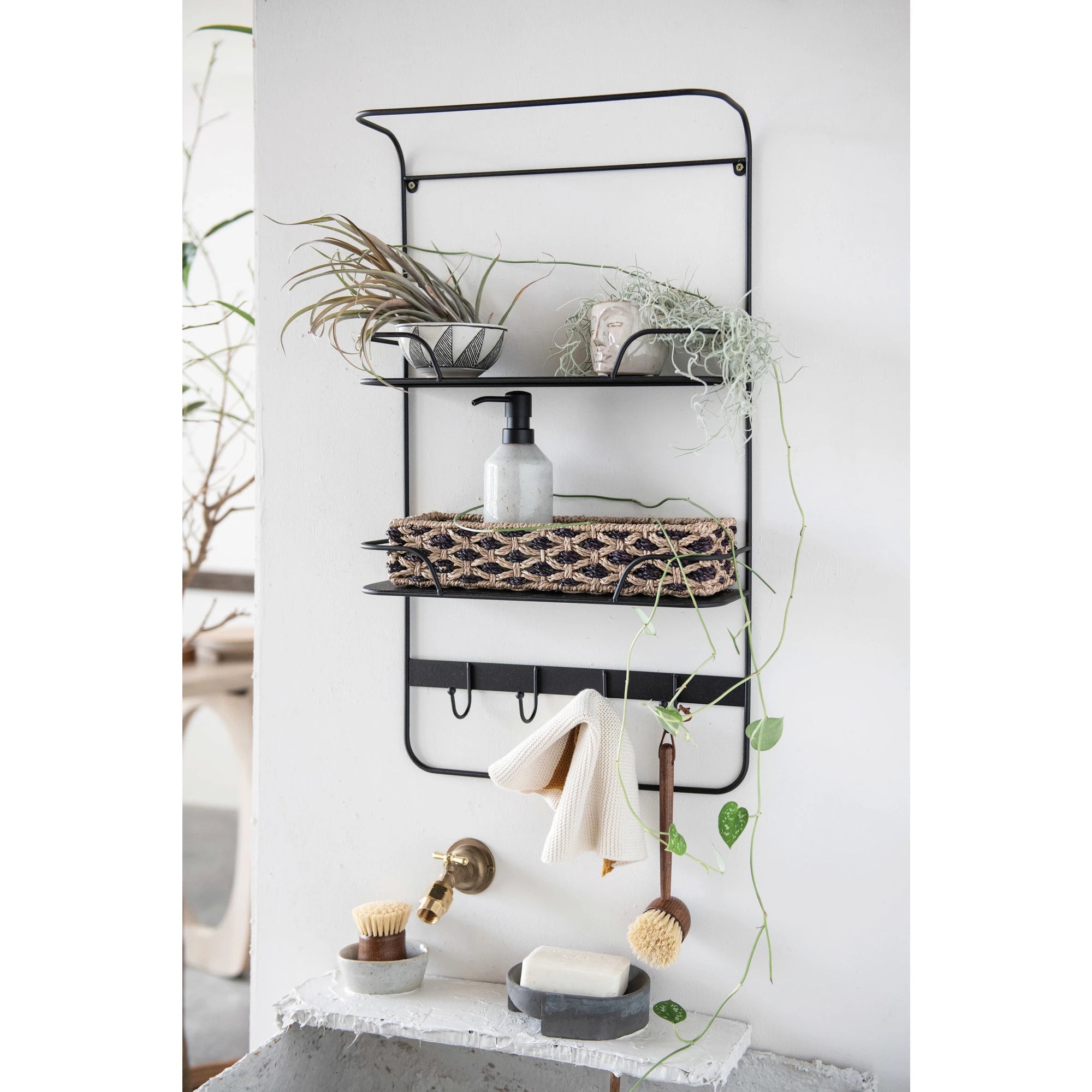 A wall-mounted, three-tiered black metal shelf displaying various bathroom items including plants, towels, and toiletries, against a white wall in a Scottsdale, Arizona bungalow featuring a Bloomingville stoneware soap dispenser.