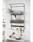 A wall-mounted, three-tiered black metal shelf displaying various bathroom items including plants, towels, and toiletries, against a white wall in a Scottsdale, Arizona bungalow featuring a Bloomingville stoneware soap dispenser.