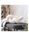 A Bloomingville Cotton Knotted Pillow in cream sits on a dark gray bench beside a blue and white knit blanket, with a white brick wall, drawing, and decorative branches in the background.