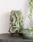The Bloomingville Green Floral Texture Vase (round stone vase) with a green palm leaf pattern sits on a gray console table beside a green glazed bowl and a tall leafy stem, set against a textured white wall.