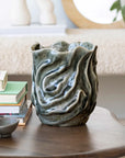 The Bloomingville Wavy Blue Stone Vase (Round Stone Vase), featuring a textured, glossy finish, sits on a wooden table beside stacked books and a small dish in a softly lit, cozy living room.