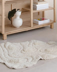 A 3'x3'8" tufted rug by Bloomingville with a wavy texture sits on a light floor in front of a wooden shelf that holds a white vase with a leaf and stacked books, creating a softly lit, minimalist scene.