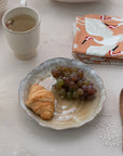 A Creative Co-op Organic Shaped Plate with a croissant and grapes sits on a light table beside tea, a wooden spoon, and a folded orange crane napkin. The plate’s reactive glaze lends subtle elegance to the breakfast scene.