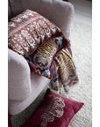 A beige armchair holds a patterned cushion, with a Bloomingville Cotton Throw/Tibetan Tigers draped over the arm. A maroon pillow with gold embroidery lies on the light carpet—a perfect boho decor accent.