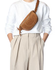 A person in a white sweater and beige pleated trousers carries the handmade Jerome Dreyfuss Lino Banana bag, a brown leather crossbody, across their chest. The photo is set against a plain white background.